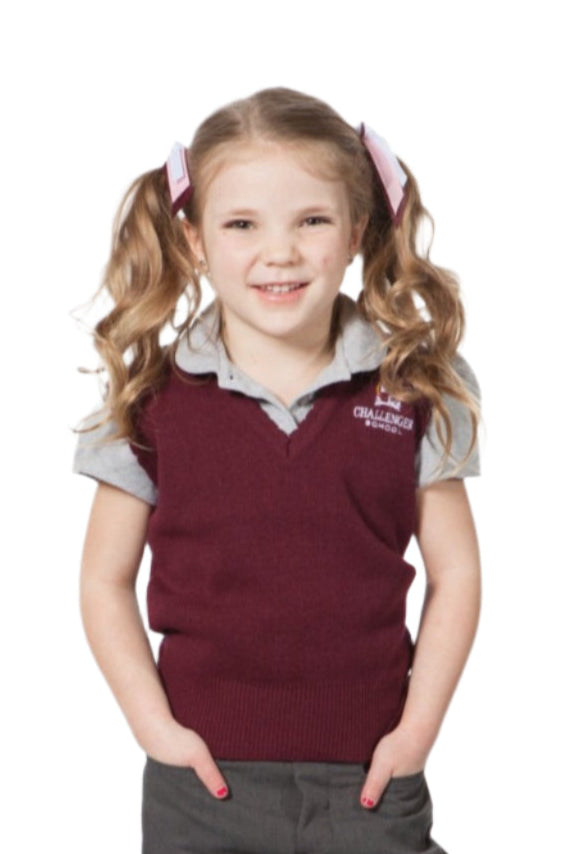A girl wearing a dark maroon sweater vest over a white shirt, paired with gray shorts and black shoes, standing with hands in pockets.